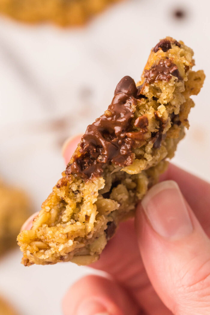 Hand holding half of a oatmeal chocolate chip cookie so you can see melted gooey chocolate chips and the texture inside the cookie.