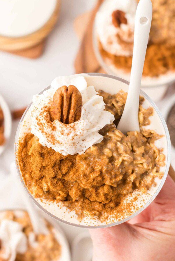 Overhead view of a hand holding a parfait glass of pumpkin overnight oats topped with whipped cream a pecan and cinnamon.