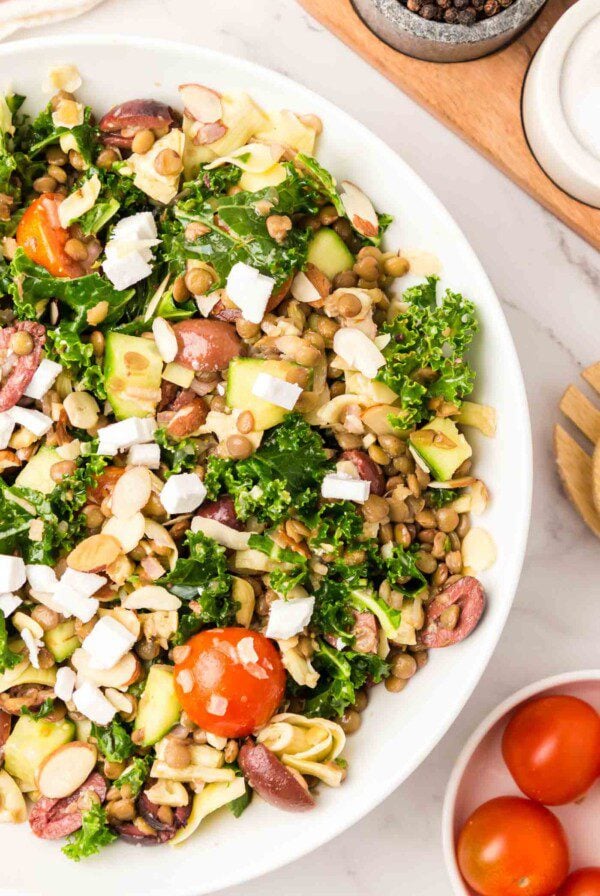 Overhead view into a large bowl of a kale salad with lentils, artichoke hearts, cucumber, feta, tomato and olives.