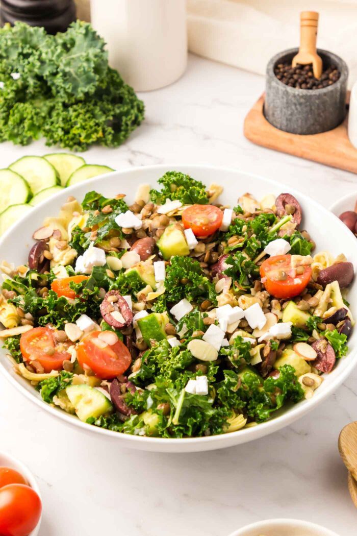 A large bowl of a kale salad with lentils, cucumbers, feta, tomatoes and olives.