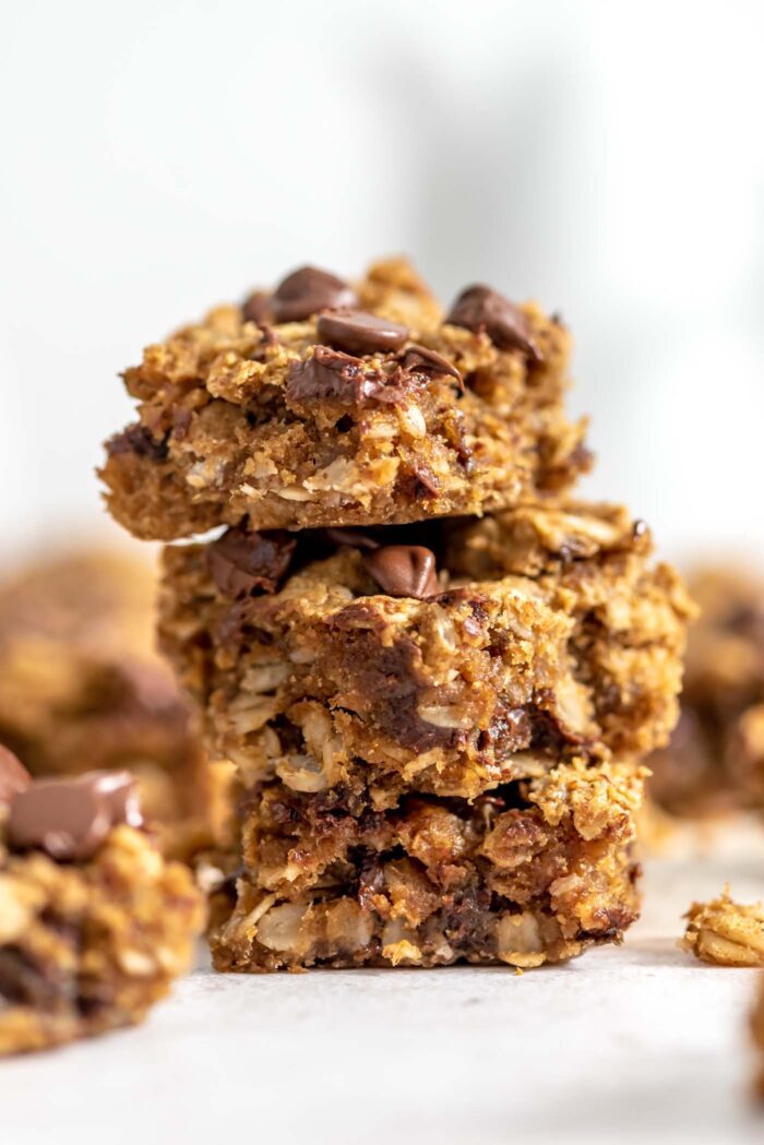 A stack of 3 peanut butter banana oatmeal bars with chocolate chips in them. More bars can be seen in the background and foreground.