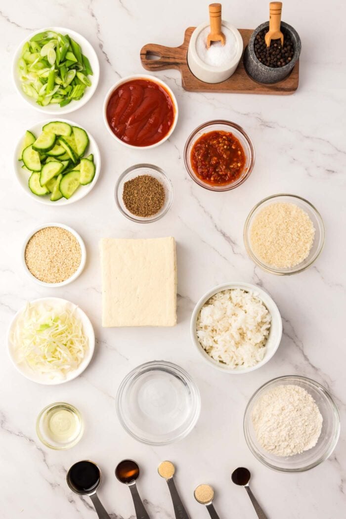 Various ingredients for making a tofu katsu recipe with rice, cabbage, cucumber and tonkatsu sauce. Each ingredient is in a small dish.