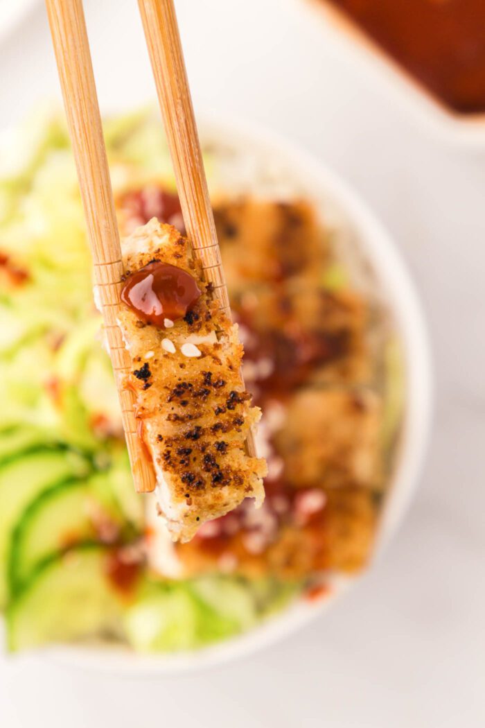 Chopsticks holding a slice of tofu katsu with sauce and sesame seeds on it.