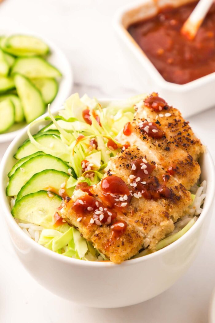 Tofu katsu cutlet cut into slices served over rice with cucumber and cabbage in a bowl with a small dish of sauce behind it.