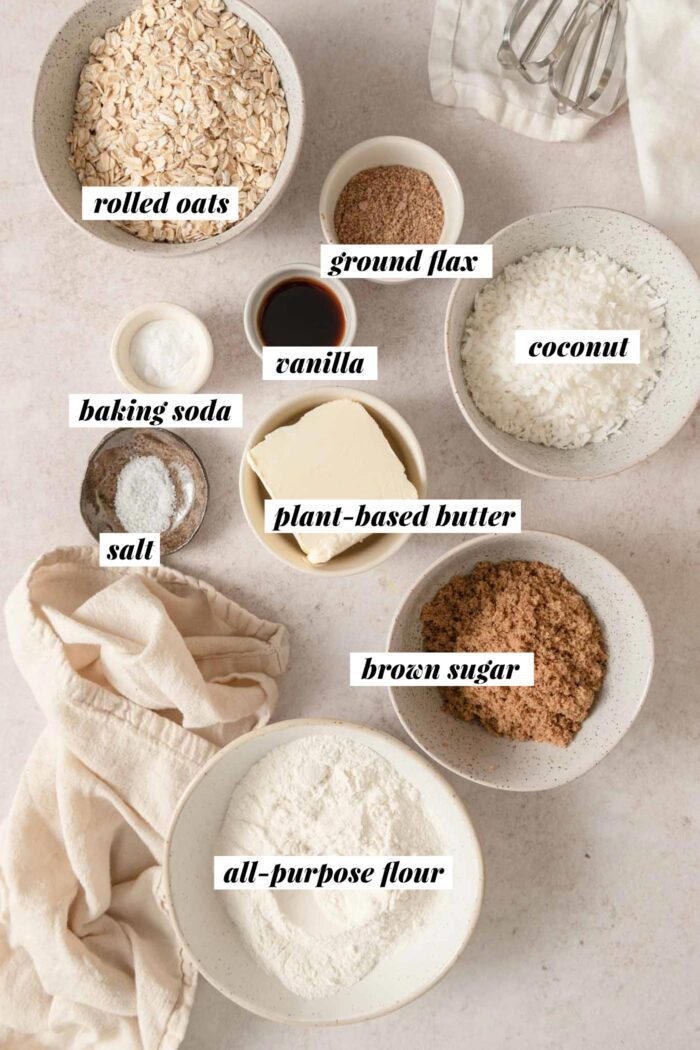 All of the ingredients for making a coconut oatmeal cookie recipe gathered in various bowls. Each ingredient is labelled with text.