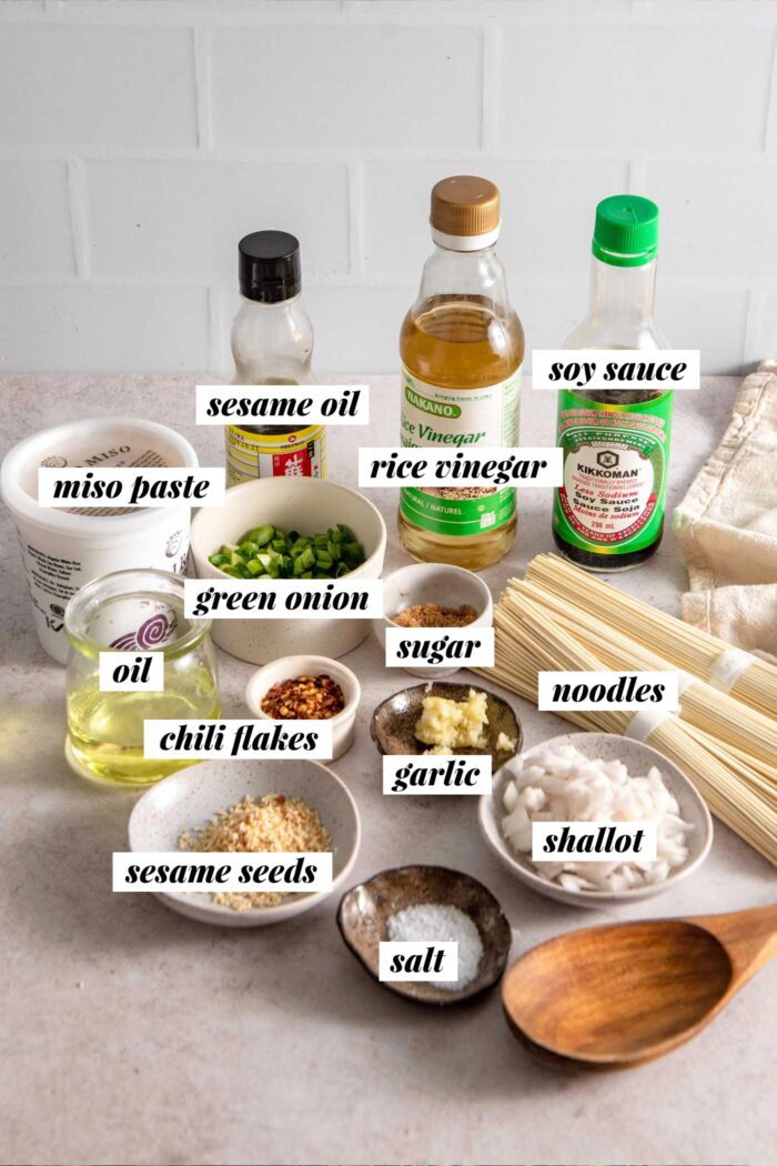 All the ingredients needed for making a garlic chili oil noodle recipe with miso paste, green onion, sesame seeds and soy sauce. Each ingredient is labelled with text.