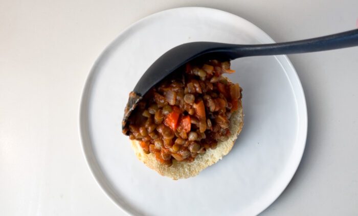 Scooping a lentil sloppy joe mixture over a toasted bun on a plate.