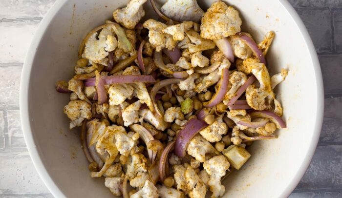 Chopped cauliflower, red onion and chickpeas mixed with curry powder and other spices in a bowl.