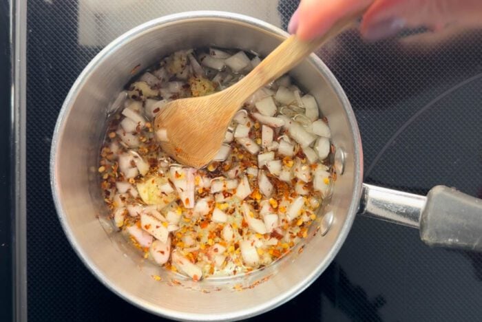 A hand stirring chopped garlic, shallot and chili flakes cooking in oil with a wooden spoon in a small pot.