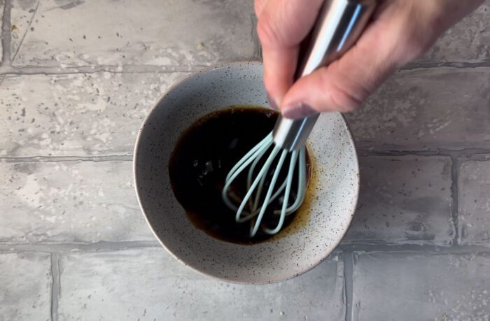 A hand mixing a dark sauce with a whisk in a small dish.