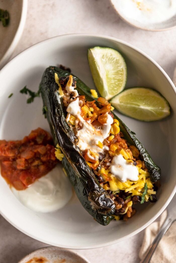 Overhead view of a poblano peppers stuffed with a vegetarian rice and bean mixture and topped with salsa and sour cream. There are some lime wedges in the bowl and small spoonfuls of salsa and sour cream.