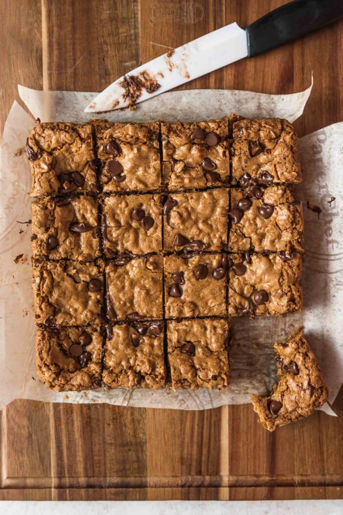 Overhead view of a batch of chocolate chip oatmeal cookie bars cut into 16 squares on a cutting board. One bar is pulled out and has a bite taken out of it.