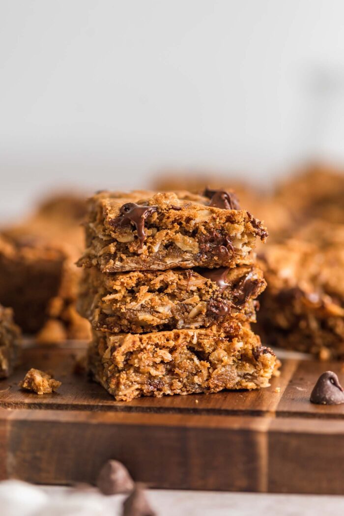 A stack of 3 oatmeal cookie bars with chocolate chips on a wooden cutting board with more bars in the background.