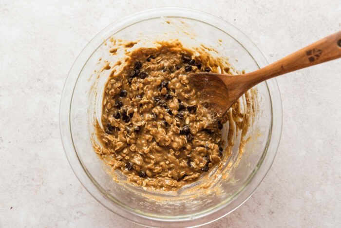 Chocolate chip oatmeal cookie bars batter in a glass mixing bowl with a wooden spoon in it.