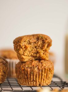 Two carrot zucchini muffins stacked on one another. The muffin on top has a bite out of it so you can see the texture and bits of grated carrot and zucchini inside.