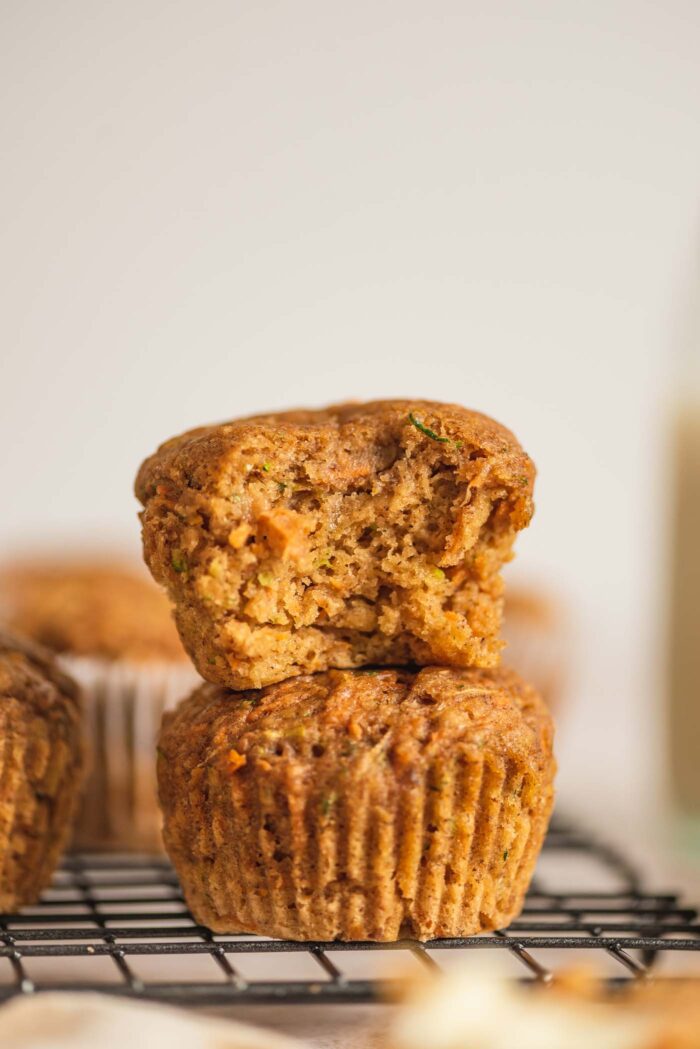 Two carrot zucchini muffins stacked on top of each other.  The muffin top has a bite so you can see the texture and bits of the grated carrot and zucchini inside.