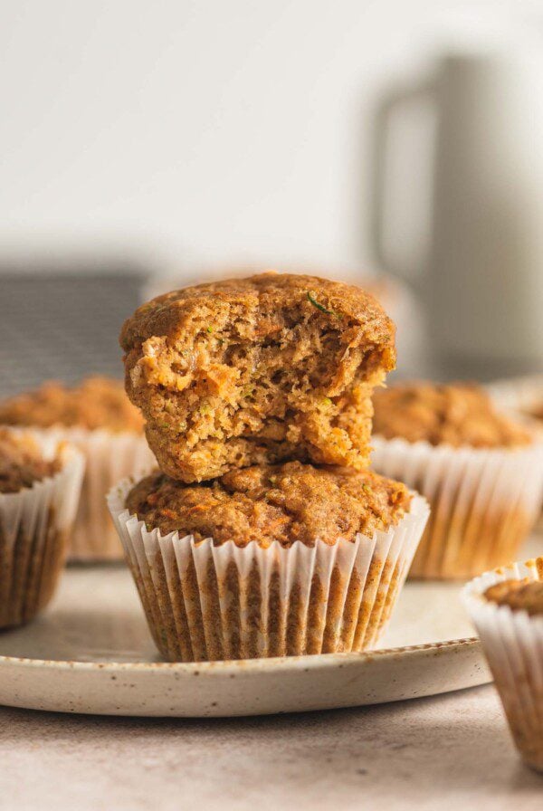 Two carrot zucchini muffins stacked on one another. The muffin on top has a bite out of it so you can see the texture and bits of grated carrot and zucchini inside.