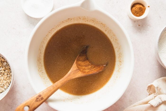 Maple syrup, brown sugar, applesauce and almond milk mixed into a brown mixture in a mixing bowl with a wooden spoon resting in it.
