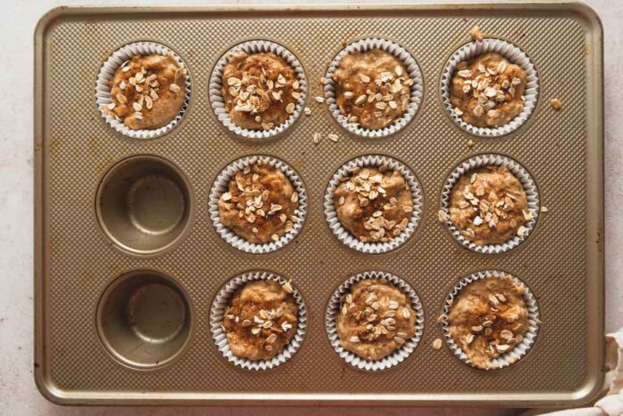 10 unbaked oatmeal muffins in paper liners in a muffin pan. Each muffin is sprinkled with rolled oats.
