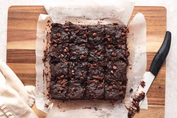 A batch of chocolate chip brownies cut into 16 squares on a wooden cutting board with a knife resting on the edge of it.