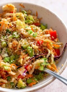 A fork resting in a small bowl of quinoa edamame salad with cabbage, green onion, red pepper, carrot and sesame seeds.