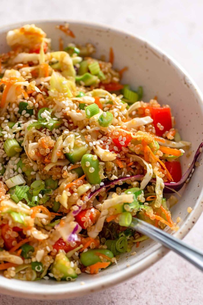 A fork resting in a small bowl of quinoa edamame salad with cabbage, green onion, red pepper, carrot and sesame seeds. 