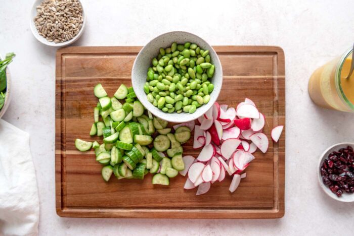 Chopped cucumbers and radishes on a cutting board with a bowl of shelled edamame beans.
