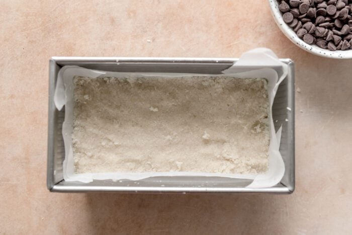 Coconut bars are pressed into a loaf pan lined with parchment paper.