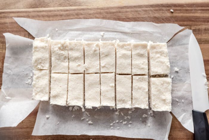 A bread pan of coconut bars is cut into 16 bite-sized bars on a cutting board.