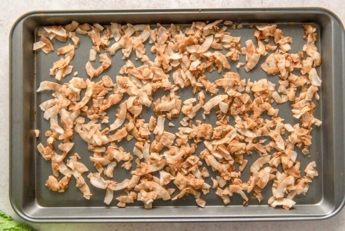 Large coconut flakes coated in a tamari mixture to make coconut bacon spread out on a baking pan.