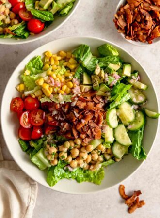 A plant-based cobb salad recipe with tomato, cucumber, corn, chickpeas and coconut bacon in a bowl with a small dish of coconut bacon beside it.