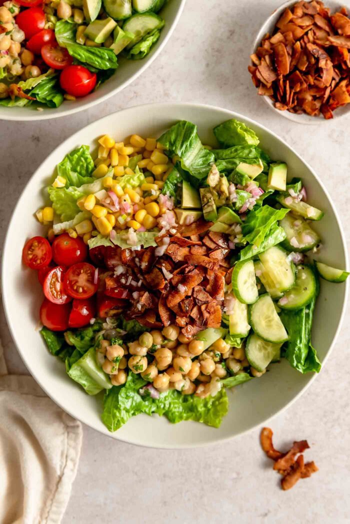 A plant-based cobb salad recipe with tomato, cucumber, corn, chickpeas and coconut bacon in a bowl with a small dish of coconut bacon beside it.