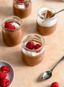 4 small glass containers of chocolate mousse topped with whipped cream and raspberries.