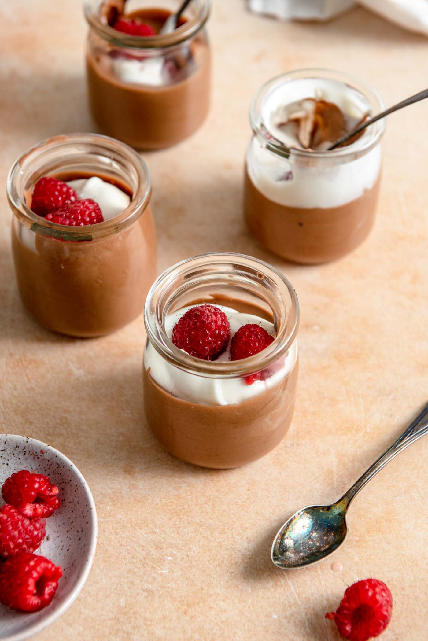 4 small glass containers of chocolate mousse topped with whipped cream and raspberries.