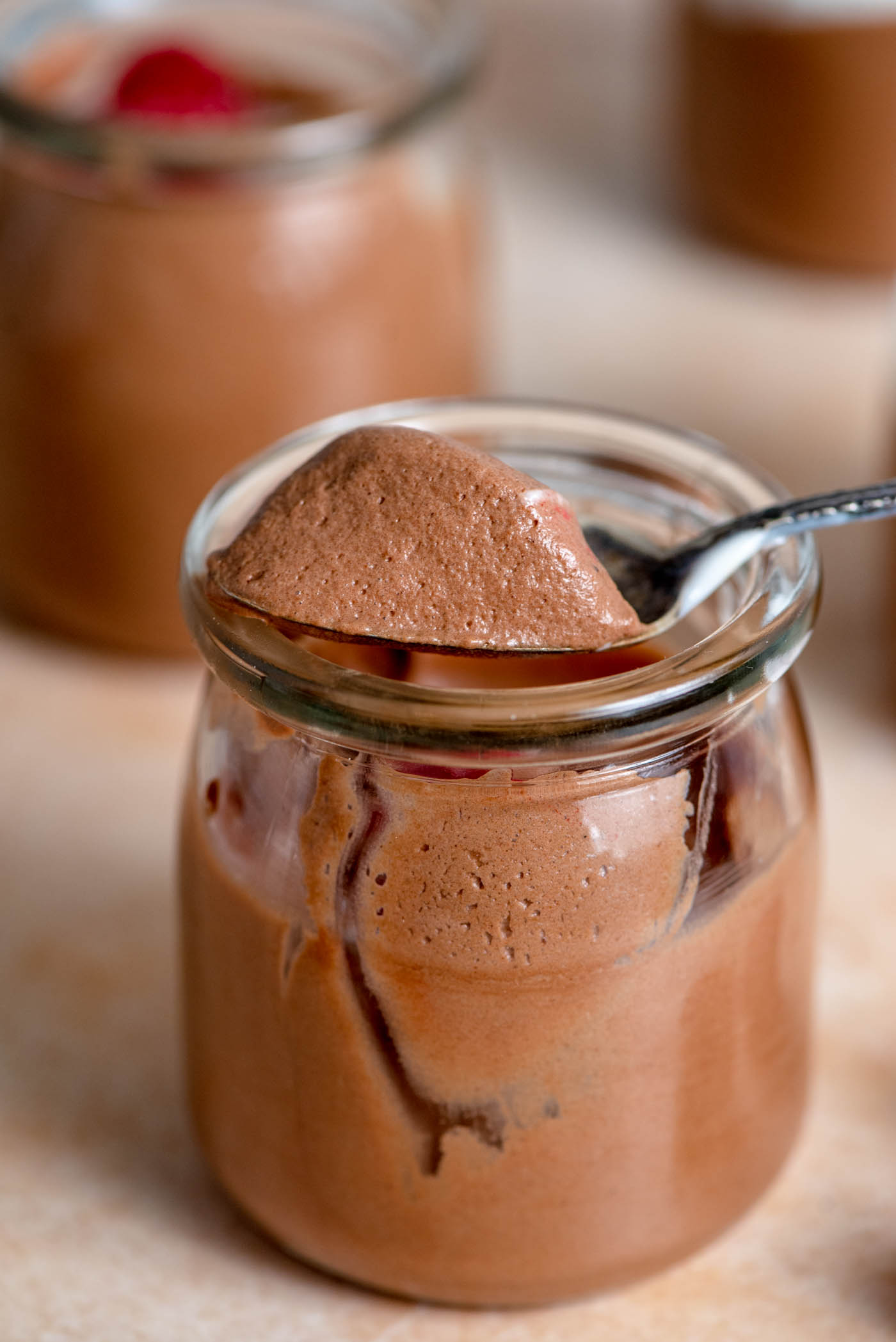 A spoonful of chocolate mousse resting on the edge of the top of a small container of mousse with raspberries and whipped cream in it.