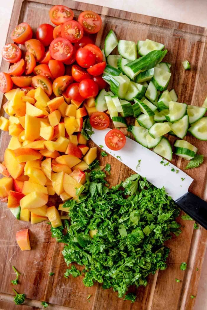 Chopped fresh herbs, nectarine, cherry tomato and cucumber on a cutting board with a knife.