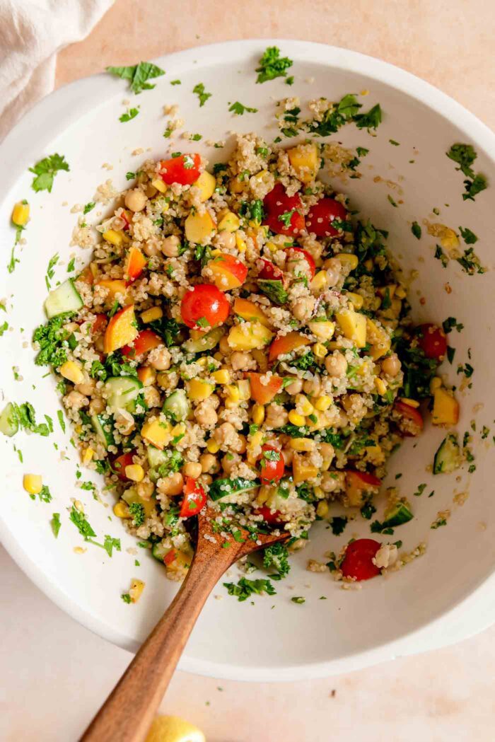 Summer quinoa salad with nectarine, chickpeas, tomato, herbs and corn in a large mixing bowl with a wooden spoon.