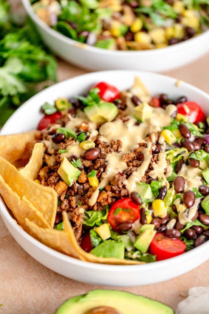A bowl of vegan taco salad with a creamy dressing drizzled over vegan taco crumbles, lettuce, tomato, avocado, black beans, red onion and corn. There are some tortilla chips on the side. 