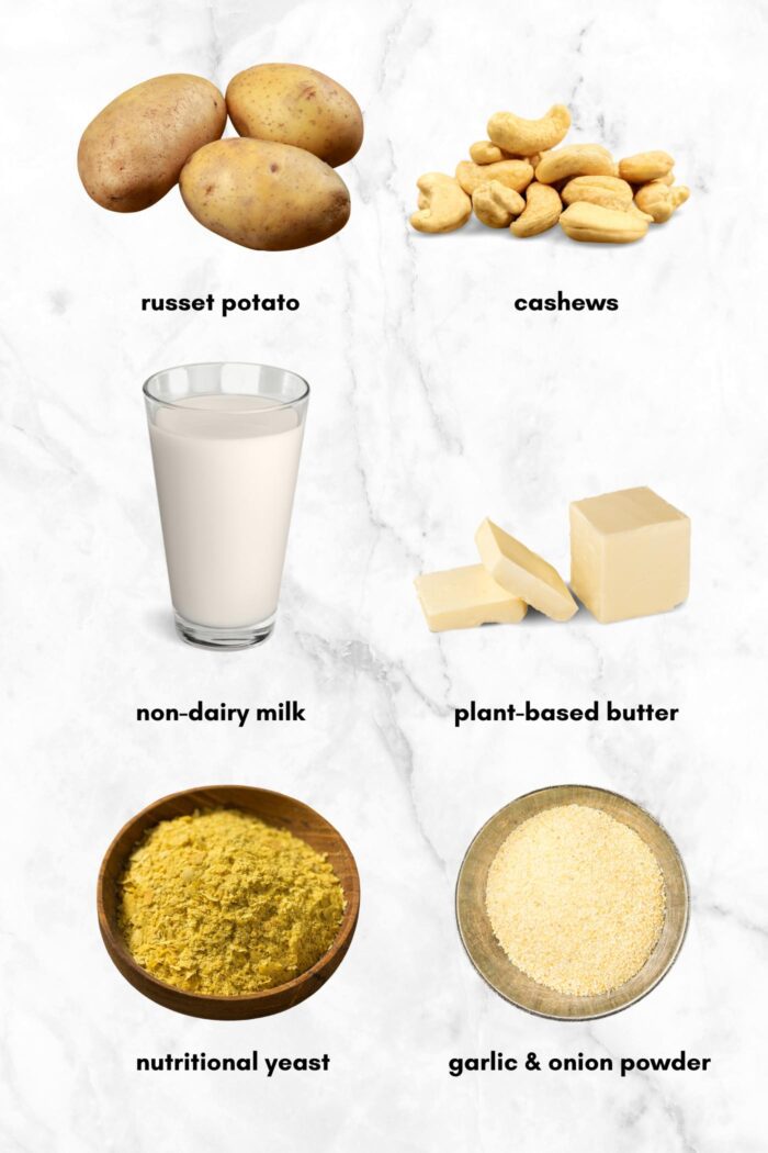 Potatoes, cashews, milk in a glass, a few small slabs of butter, bowls of nutritional yeast and garlic powder on a marble background, each labelled with text.