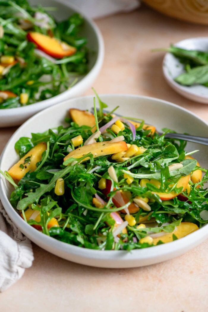 Two shallow bowls of arugula salad with peach, corn, almonds and shallots.