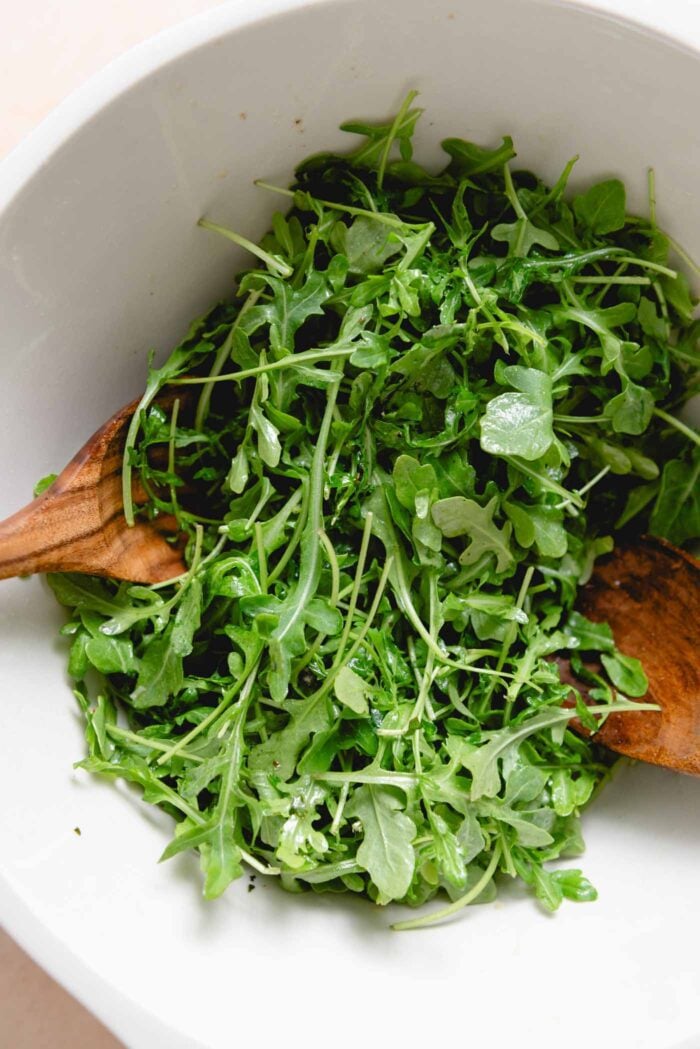 Arugula in vinaigrette in a mixing bowl with salad tongs.
