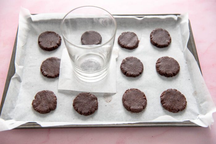 A glass sitting on a piece of parchment paper on one small, round cookie of a tray of 12 cookies. The glass is being used to flatten the cookies.
