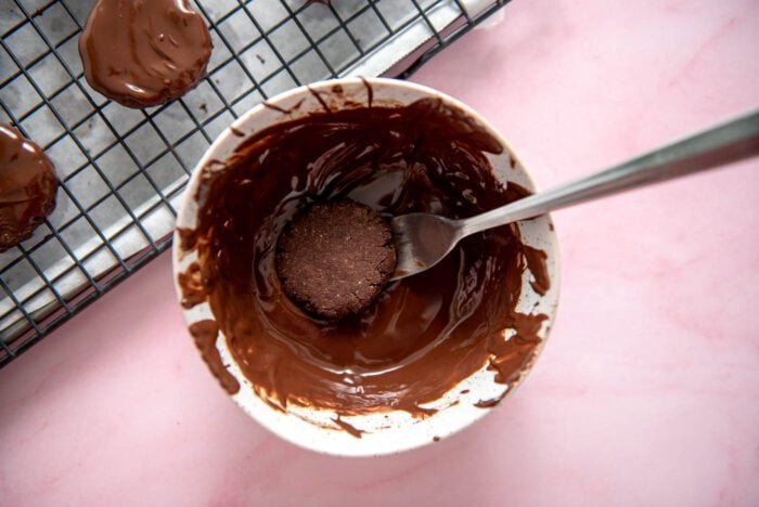 A small chocolate cookie being dipped into a bowl of melted chocolate using a fork.