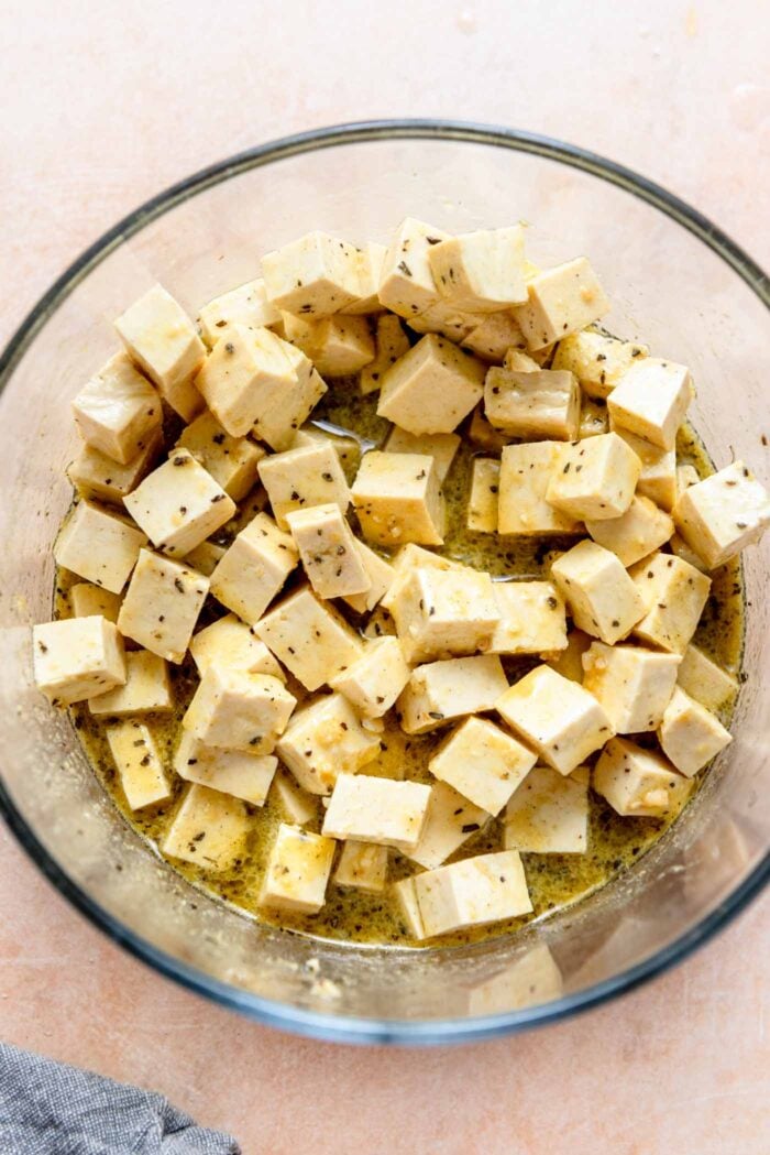 Cubes of tofu marinating in a herb, lemon and olive oil marinade in a glass bowl.
