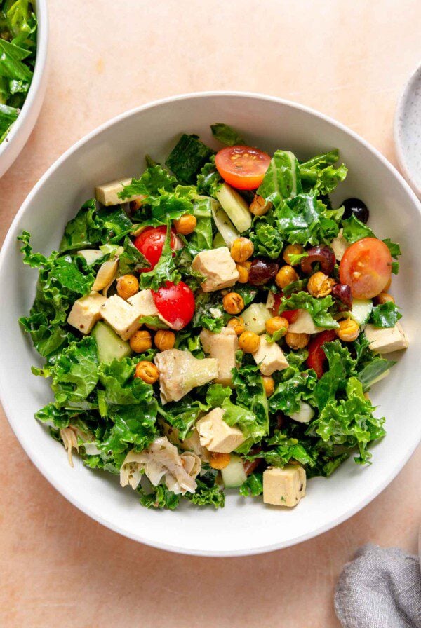 Bowl of kale salad with chopped tomatoes, artichoke hearts, cucumber, roasted chickpeas, cubes of tofu feta and chopped olives.