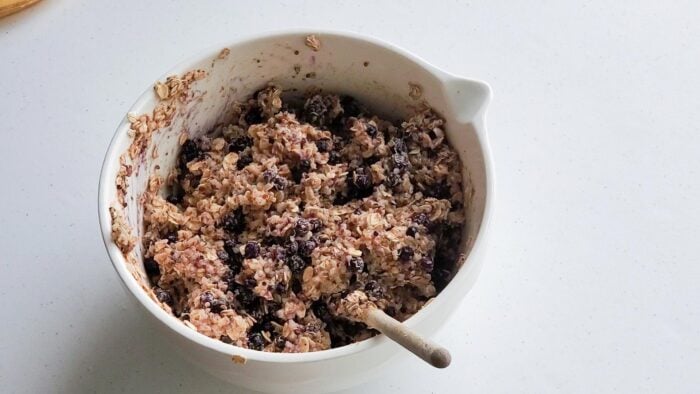 Thick batter with blueberries in it in a large mixing bowl with a spoon resting in it.