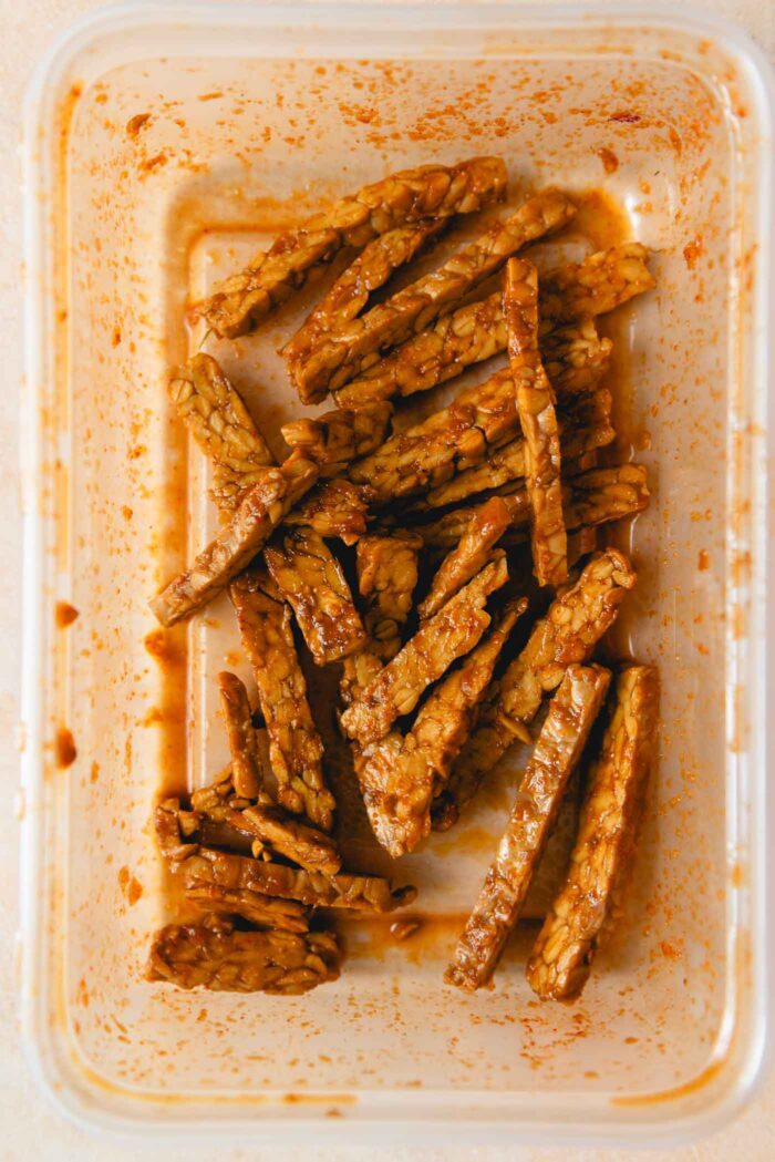 Slices of tempeh marinating in a sauce in a Tupperwear container.