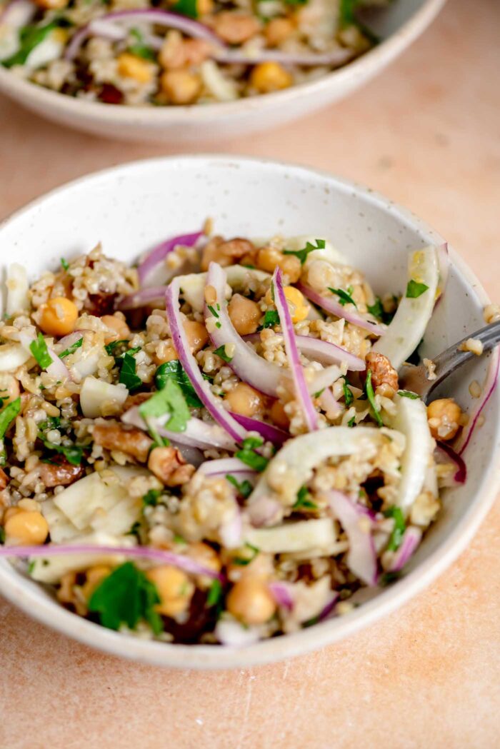 Freekeh salad with red onion, fennel, chickpeas, dates, walnuts and fresh herbs in a serving bowl with a fork.