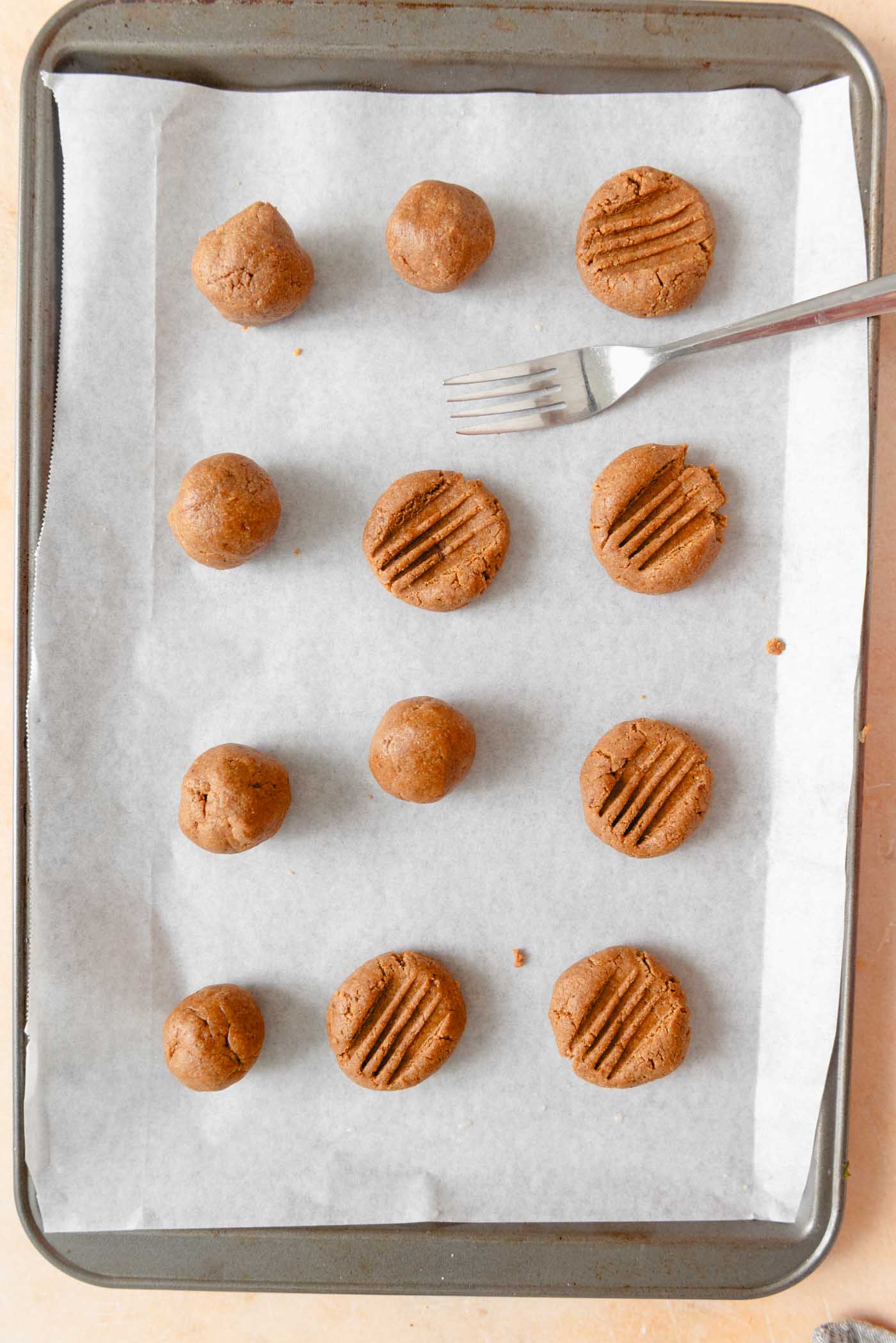 12 peanut butter cookie dough balls being pressed down by a fork on a baking sheet.