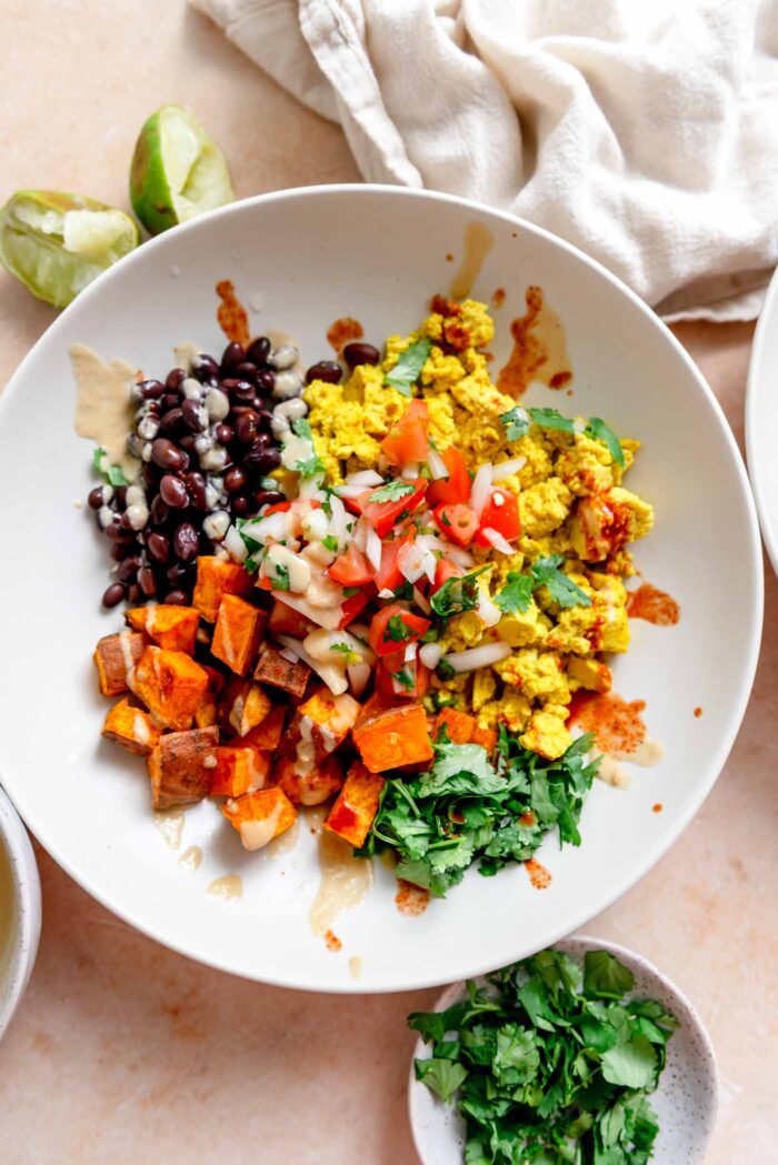 Breakfast burrito bowl with cilantro, tofu scramble, black beans, roasted sweet potato, chipotle sauce and hot sauce drizzled over top.